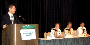 Photo of speakers before an audience, with the man at the left standing before a lectern.